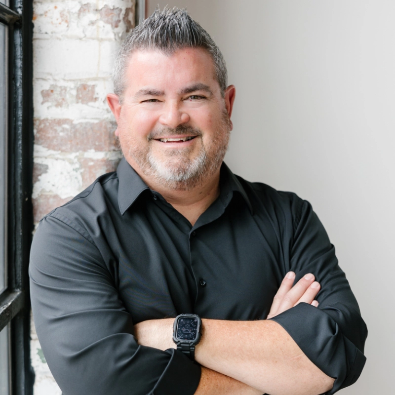 Kent Vincent, with gray hair and a beard, wearing a black shirt and smartwatch, stands with his arms crossed, smiling in front of a partially exposed brick wall.