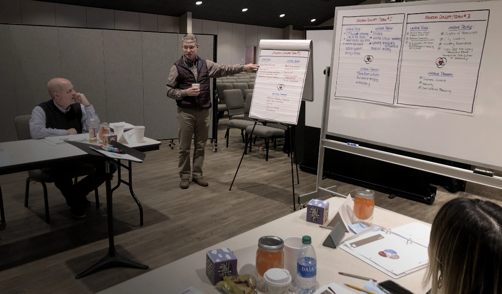 A man presents to people in a conference room, pointing at a flip chart with handwritten notes. Tables with beverages and papers are in the foreground, indicative of an intense brainstorming session about church consulting services. A large whiteboard with notes fills the background.