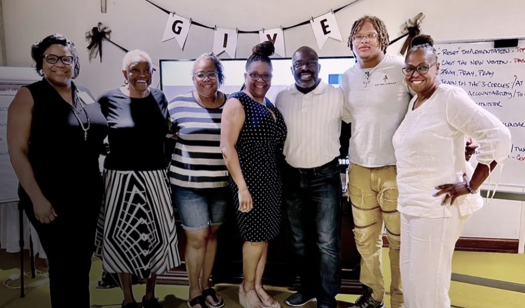 A group of seven people stand smiling in a room with a banner that reads "GIVE" above them. They are in front of a large screen and a whiteboard, appearing to attend an event or meeting hosted by church fundraising consultants.
