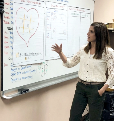 A woman stands next to a whiteboard with an illustrated heart and various notes, pointing at a section while explaining missional communities.