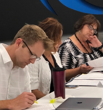 Three people sit at a table concentrating on documents. One holds a highlighter, another rests their head on their hand while reading, and a closed laptop is visible on the table.