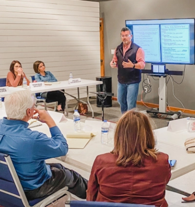 A church consultant stands and speaks in front of a screen displaying text, while several people sit at tables listening and taking notes in a meeting room.